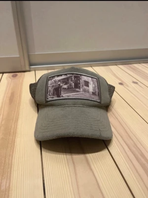 Grå keps  meshkeps med vintage-tryck - Säljer en grå keps med mesh på sidorna och bak, samt ett vintage-inspirerat tryck framtill. Justerbar storlek med plastspänne och snygg detalj i form av en metallogo på sidan. Perfekt för dig som gillar streetwear och retrostil. Ganska sliten men funkar bra, därav priset, bara att skriva om ni undrar något. Mvh Nils 