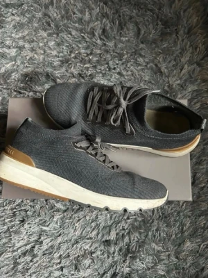 Grå sneakers från Brunello Cucinelli - Säljer ett par stilrena grå sneakers från Brunello Cucinelli i storlek 43. Skorna har ovandel i stickad textil, vita och bruna detaljer på sulan samt snörning. Perfekta för dig som gillar exklusiva och bekväma sneakers med modern design. Pytteliten slitning/hål på högra skon som inte märks vid användning, nypris runt 10k