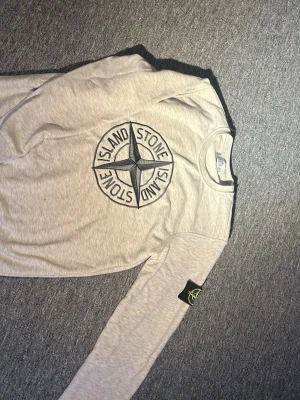 Ljusgrå långärmad tröja Stone Island - Säljer en ljusgrå långärmad tröja från Stone Island med stort broderat logomärke på bröstet och klassisk patch på ärmen. Tröjan har rund hals och är tillverkad i mjuk bomull, perfekt för dig som gillar streetwear och snygga detaljer.