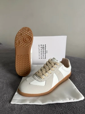 Maison Margiela skor  - Maison Margiela Replica sneakers i vitt och beige med klassisk gummisula. Ovandelen är i en mix av skinn och mocka, med beige snörning och rund tå. Snygg minimalistisk design och diskret logga på plösen. Perfekta för dig som gillar stilrena och trendiga sneakers.