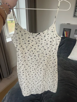 Vit blommig klänning Brandy Melville - Supersöt vit kortklänning från Brandy Melville med tunna axelband och svart blommigt mönster. Klänningen har en enkel, rak passform och är tillverkad i mjuk bomull som känns skön mot huden. Perfekt för varma dagar och enkel att styla med sneakers eller sandaler.