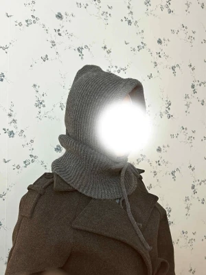 Mörkgrå stickad Balaklava  - Säljer en mörkgrå stickade Balaklava med  och dragsko. Västen har en ribbad struktur och täcker både axlar och hals, vilket ger en cool och mysig look. Perfekt att slänga över en jacka för extra värme och stil.