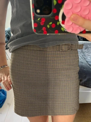 Rutig beige kortkjol med bälte - Säljer en snygg kortkjol i beige och svart med klassiskt rutigt mönster. Kjolen har en rak passform och ett matchande bälte i midjan för extra stilpoäng. Perfekt att styla med en enkel t-shirt eller stickad tröja. Passar dig som gillar tidlös och trendig look.