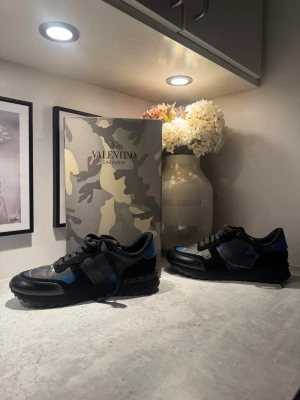 Valentino Rockrunner svart/blå - Säljer ett par Valentino Rockrunner sneakers i svart och blått med coolt kamouflagemönster. Skorna har snörning, platt sula och rund tå. Materialmix av skinn och mocka med detaljer i syntet. Kommer med originalkartong och dustbag. Använda max 2 ggr. Pris kan diskuteras hör gärna av dig!!