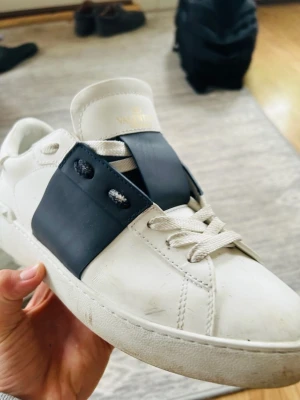 Valentino Open sneakers vit/blå - Säljer ett par Valentino Open sneakers i vitt skinn med svart band över snörningen. Klassisk låg modell med rund tå, vita snören och nitar på hälen. Snygg kontrast mellan det vita och svarta, perfekt för dig som gillar stilrena men ändå unika skor.