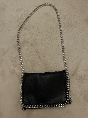 Svart axelväska med kedja från Tiamo - Cool svart axelväska från Tiamo i skinnimitation med grov silverfärgad kedja som axelrem och runt kanterna. Väskan har magnetknäppning, innerfack med dragkedja och ett ytterfack med dragkedja på baksidan. Perfekt storlek för det viktigaste och ger en edgy vibe.