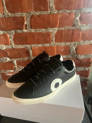 Oliver Cabell svarta sneakers med vit detalj - Snygga svarta sneakers från Oliver Cabell med vit sula och vit cirkeldetalj på sidan. Skorna har snörning med svarta skosnören där märket står tryckt. Ovandelen är i skinn och hälen har en vit panel med logga. Perfekt för dig som gillar stilrena och moderna sneakers. 