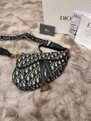 Dior Saddle axelväska i blått mönster - Säljer en ikonisk Dior Saddle axelväska med blått och vitt mönster, detaljerad med guldfärgade metalldelar och en bred axelrem med broderade inslag. Väskan har en unik sadelform och är tillverkad i bomull med skinn-detaljer. Perfekt statement piece med klassisk Dior-logga.