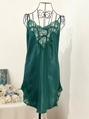 Vintage grön slipklänning från Victoria's Secret - Snygg grön slipklänning i satin med tunna axelband och dekorativ spets vid halsringning och nederkant. Vintage Gold Tag av Victoria's Secret. Storlek: M, passar 38-40. Dockans storlek: 34.