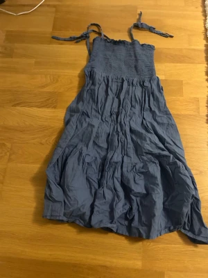 Blå kort klänning - Säljer en blå klänning från Ellos i storlek 34/36. Klänningen har knytband vid axlarna och smock upptill, vilket ger en skön och avslappnad passform. Materialet är mjukt och lätt, perfekt för varmare dagar. Den har en volangig och rynkad nederdel som ger extra rörelse.