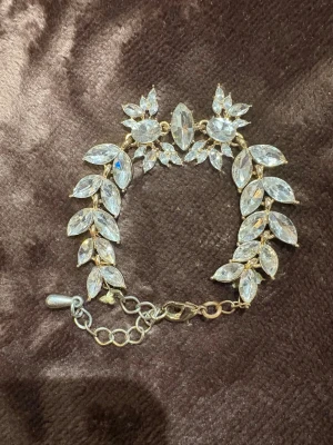 Guldig armlänk med kristallblad - Säljer ett glittrigt armband i guldmetall med stora, klara kristaller formade som blad och blommor. Armbandet har justerbar kedja och stängs med karbinhake. Perfekt för att ge outfiten extra bling och wow-känsla.