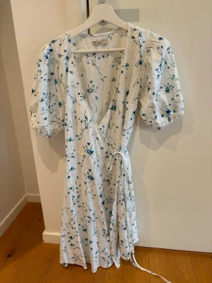 Vit blommig omlottklänning  - Superfin vit omlottklänning  med blått blommigt mönster. Klänningen har puffiga korta ärmar, v-ringning och knyt i midjan. Tillverkad i linne, perfekt för varma dagar och har en luftig känsla.