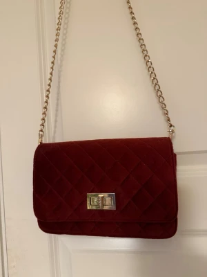 Vinröd axelväska med kedja - En vinröd axelväska med guldfärgad kedja som axelrem. Väskan har en rektangulär form och ett snyggt metallspänne framtill. Materialet är mjuk mocka som ger en lyxig känsla. Perfekt för dig som vill ha en stilren och trendig väska.