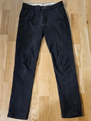 Svarta chinos från Jack & Jones slim fit - Svarta chinos från Jack & Jones Pants Studio i slim fit-modell. Byxorna har klassisk design med framfickor, bakfickor med knapp och bälteshällor. Materialet är bomull och de har dragkedja i gylfen. Perfekt för dig som gillar stilrena och bekväma byxor.