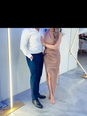 Beige långklänning med slits - Säljer en elegant beige långklänning med hög hals och ärmlös design. Klänningen har en snygg drapering i midjan och en hög slits framtill som ger en modern och stilren look. Perfekt för dig som vill sticka ut på festen!