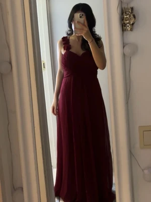 Oanvänd Vinröd långklänning med axelblommor - Säljer en vinröd långklänning med v-ringad omlottliv och dekorativa blommor på ena axelbandet. Klänningen har ett flytande fall och är ärmlös, perfekt för bal eller fest. Materialet känns lätt och elegant.😍❤️