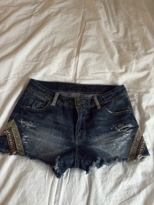 Mörkblå jeansshorts från Stradivarius - Säljer ett par mörkblå jeansshorts från Stradivarius med slitna detaljer och fransig kant. Shortsen har broderade mönster i färg på sidorna och klassisk femficksdesign💕