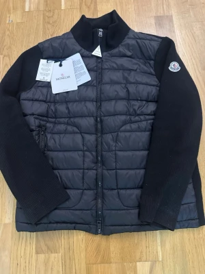 Moncler Cardigan  - Säljer en svart cardigan  från Moncler med quiltad front och stickade ärmar. Jackan har dragkedja framtill, Moncler-logga på ärmen och ribbad krage. Perfekt för dig som vill ha en stilren och exklusiv look.
