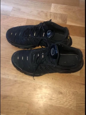 Nike Tn - Svarta Nike tn med mesh- och syntetmaterial, ikoniska vågiga detaljer och synlig Air-dämpning i sulan. Skorna har snörning och en platt sula med mönstrad undersida. Swoosh-logga på sidan och TN-märke under sulan.