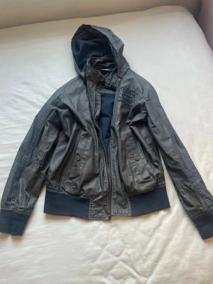 Grå/mörblå vindjacka med huva från Out Wear - Snygg mörkblå vindjacka från Out Wear med broderad text på bröstet och praktisk huva. Jackan har dragkedja framtill, två fickor med tryckknappar och ribbade muddar vid ärmslut och nederkant. Perfekt för blåsiga dagar och chill stil. Barnjacka i storlek 158