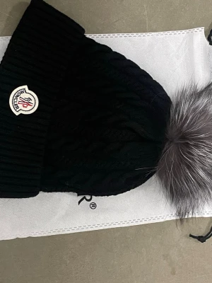 Svart Moncler mössa med pälsboll - Snygg svart mössa från Moncler med grovstickad struktur och bred ribbad kant. Mössan har en stor fluffig pälsboll i grå toner på toppen och Moncler-logga framtill. Perfekt accessoar för att höja vinterlooken.