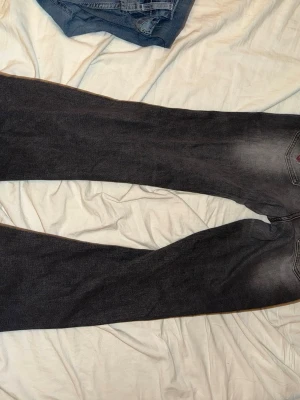 Grå bootcut jeans med strassdetalj - Snygga grå jeans med bootcut passform och coolt strassmönster i rött och silver på bakfickan. Jeansen har klassisk femficksdesign och är tillverkade i mjukt bomullsmaterial med lite stretch. Perfekt för dig som gillar detaljer och vill sticka ut.