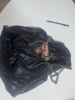 Svart axelväska med kedjedetaljer - Säljer en svart axelväska med glansig yta och grova silverfärgade kedjor längs kanterna. Insidan har leopardmönstrat foder och en praktisk dragkedja. Perfekt för dig som gillar edgy och cool stil. Väskan är rymlig och har en mjuk form.