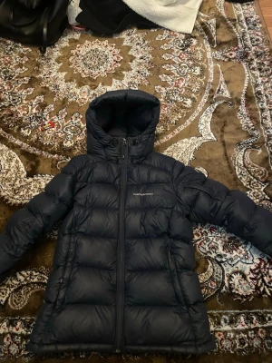 Mörkblå Peak Performance dunjacka - Säljer en mörkblå dunjacka från Peak Performance med huva och dragkedja framtill. Jackan har quiltad design och långa ärmar, perfekt för kalla dagar. Snygg och funktionell med stilren look och logga på bröstet.