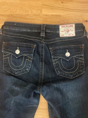 True Religion Becca Mid Rise Bootcut Jeans - Säljer ett par mörkblå jeans från True Religion, modell Becca Mid Rise Bootcut. Jeansen har klassiska kontrastsömmar, snygga fickdetaljer med lock och silverfärgade knappar. Bootcut-snittet ger en schysst siluett och de är tillverkade i jeansmaterial.