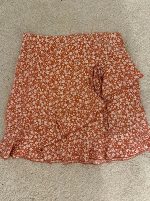 Blommig omlottkjol med volang - Säljer en kort omlottkjol i rosa med vita blommor. Kjolen har volangkant längst ner och knyt i sidan för en snygg passform. Perfekt för dig som gillar färgglada och somriga plagg med lite extra detaljer.