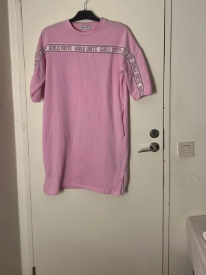 Rosa långklänning  - Pris kan diskuteras och tvätt ingår innan leverans 💗Ljusrosa lång klänning från FB Sister med texten 'GIRLS UNITE' i vitt och lila längs bröstet och ärmarna. Klänningen har korta ärmar, rund halsringning och är gjord i mjuk bomull. Perfekt för en chill och avslappnad stil.