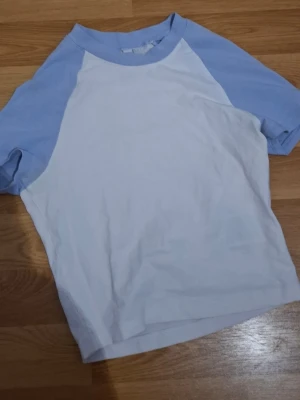 t-shirt - Säljer en basic t-shirt från H&M Divided i storlek XS. T-shirten har vit kropp och ljusblå raglanärmar samt krage. Tillverkad i mjuk bomull, perfekt för chill dagar. Enkel och stilren design som funkar till jeans eller shorts.