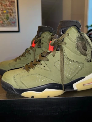 Nike Air Jordan 6 Travis Scott Olive - Säljer ett par Nike Air Jordan 6 Travis Scott i olivgrönt mocka med svarta och beige detaljer, röda accenter och ficka på sidan. Ikoniska broderade loggor bak och cool retro-vibe. Perfekta för dig som vill sticka ut med limited edition sneakers.