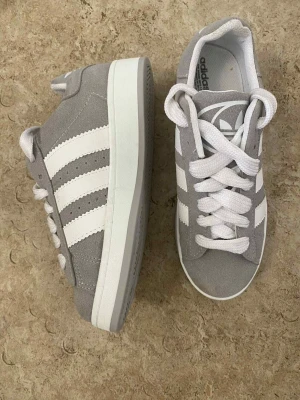 Grå Adidas Gazelle sneakers - Säljer ett par grå Adidas Gazelle sneakers med klassiska vita ränder på sidorna och vit platt sula. Skorna har rund tå, snörning och är tillverkade i mocka med syntetdetaljer. Perfekta för dig som gillar stilrena och tidlösa sneakers.
