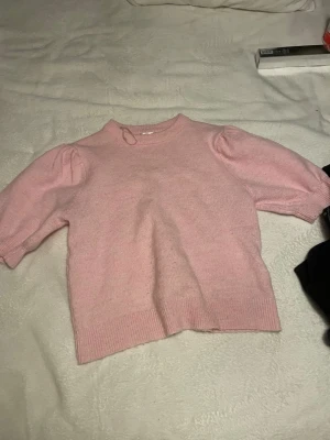 Ljusrosa stickad tröja med puffärm - Säljer en ljusrosa stickad tröja med puffade korta ärmar och rund hals. Tröjan har ribbade muddar vid ärmslut och nederkant, vilket ger en snygg och modern look. Perfekt för dig som gillar mjuka pasteller och enkel stil.