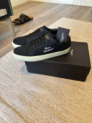 Svarta Saint Laurent sneakers - Svarta sneakers från Saint Laurent med vit text på sidan och kontrasterande vit sula. Skorna har rund tå, snörning och detaljer i skinn och mocka. Snygg och stilren design som passar till streetwear och casual outfits. Skorna är alldrig andvända och är i helt nytt skick. Perfekta vinter skorn för dig