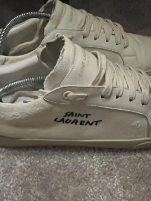 Saint Laurent vita sneakers med snörning - Säljer ett par stilrena vita sneakers från Saint Laurent med handskriven logga på sidan. Skorna har rund tå, platt sula och klassisk snörning. Materialet är en mix av canvas och skinn, vilket ger en cool och avslappnad vibe. Perfekta för dig som gillar minimalistisk design.