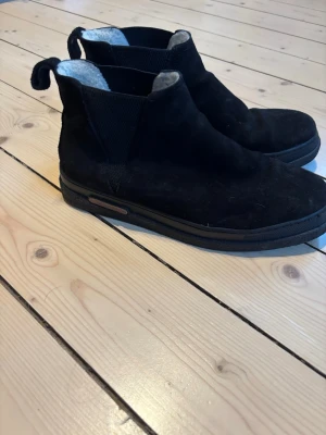 Svarta chelsea boots från Gant - Säljer ett par svarta chelsea boots från Gant i mocka med elastiska paneler på sidorna och mjukt foder på insidan. Skorna har platt sula och rund tå, perfekta för höst och vinter. Klassisk och stilren design som funkar till många outfits.
