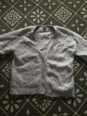 Ljuslila mjuk kofta - Supermjuk och fluffig ljuslila kofta från Lindex med lång ärm och v-ringad hals. Koftan har tre matchande knappar framtill och ribbade muddar vid ärmslut och nederkant. Syns inte så bra men den är lila och vit rutig. Den är inte lika mjuk längre pågrund av många tvättar😓