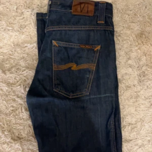 Mörkblå jeans från Nudie Jeans Co - Säljer ett par mörkblå jeans från Nudie Jeans Co i modellen med klassisk bakficka och orange sömmar. Jeansen är tillverkade av 100% ekologisk bomull och har en snygg, tidlös look. Perfekta för dig som gillar stilrena och hållbara jeans.