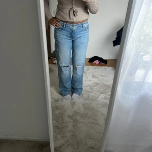 Ljusblå slitna jeans med raka ben - Säljer ett par ljusblå jeans med raka ben och slitningar vid knäna. Jeansen har klassisk femficksdesign och låg midja. Perfekta för en avslappnad och trendig look.