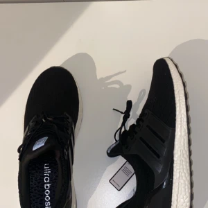 Adidas Ultraboost svarta sneakers - Svarta Adidas Ultraboost sneakers med vit, responsiv sula och klassiska tre ränder på sidan. Ovandelen är i stickad syntet för en skön passform och ventilerande känsla. Skorna har snörning och en platt sula som ger bra komfort till vardags. Skorna är helt nya och oanvända.