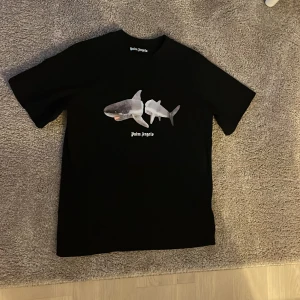 Svart Palm Angels t-shirt med hajmotiv - Svart t-shirt från Palm Angels med tryck av en haj på framsidan och stor vit Palm Angels-logga på ryggen. Klassisk passform och rund hals. Tillverkad i mjuk bomull, perfekt för en streetwear-look. Oanvänd (nyskick)
