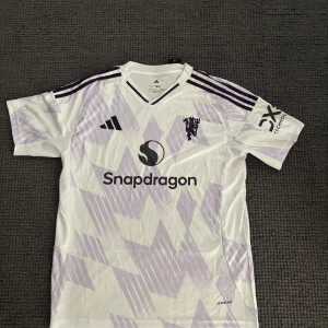 Manchester United bortatröja Adidas L - Säljer en Manchester United bortatröja från Adidas i storlek L. Tröjan är vit med ljuslila grafiskt mönster, har korta ärmar och klassiska Adidas-ränder på axlarna. Materialet är lätt och andas, perfekt för fotboll eller träning. Snapdragon-logga på bröstet.
