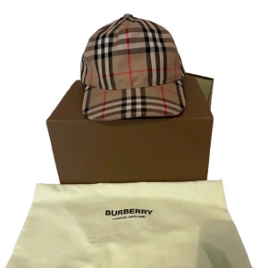 Burberry keps - Snygg keps från Burberry med klassiskt rutigt mönster i beige, svart, vitt och rött. Justerbar baktill med svart plastspänne och tydlig Burberry London England-logga. Perfekt accessoar för dig som vill sticka ut med lyxig streetstyle.