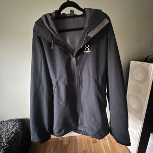 Svart skal och regnjacka från Haglöfs - Svart skal och regnjacka från Haglöfs med huva och hel dragkedja framtill. Jackan har minimalistisk design, två sidofickor och diskret logga på bröstet. Perfekt för blöta dagar och enkel att matcha med allt. Storlek L. Köpt på Intersport. Endast använd ett fåtal gång. 