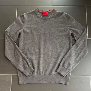 Grå stickad tröja från Hugo Boss - Snygg grå stickad tröja från Hugo Boss med rund halsringning och diskret logga vid fållen. Tröjan har långa ärmar och ribbade muddar vid ärmslut och nederkant. Storlek M men sitter som S. Har en liten gul fläck, knappt synlig 