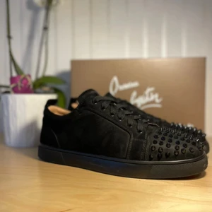 Christian Louboutin svarta sneakers med nitar - Svarta sneakers från Christian Louboutin i mocka med coola svarta nitar på tån och klassisk röd sula med logga. Skorna har snörning och platt sula, samt en rund tå. Perfekt för dig som vill sticka ut med en lyxig och edgy stil.