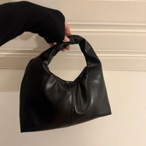 Svart handväska i skinnimitation - Trendig svart handväska i mjuk skinnimitation med minimalistisk design och rundad form. Väskan har ett kort handtag och stängs med magnetknapp. Perfekt storlek för det viktigaste och passar till alla outfits.