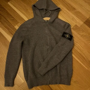  Stone Island cardigan - Säljer en grå stickad cardigan från Stone Island med dragkedja och klassisk logga på ärmen. Tröjan har huva, ribbad struktur och långa ärmar. Perfekt för dig som gillar streetwear och vill ha något både snyggt och bekvämt.